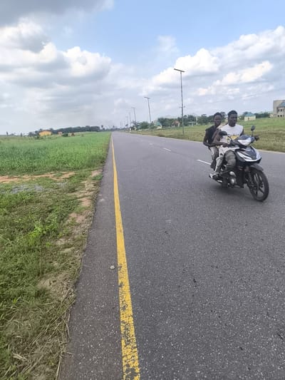 Two young men are depicted riding a black and silver motorcycle on a paved road in Abraka, Nigeria. The driver, wearing a white sleeveless shirt with the word "Fashion" visible on it and light-colored pants, steers the motorcycle. Behind him, the passenger wears a dark sleeveless shirt and dark trousers. Both riders are looking forward, and the motorcycle appears to be in motion.
The road features a faded yellow solid line marking its edge on the left and a faint dashed white line visible further down the center. On both sides of the asphalt, vast stretches of vibrant green grass and wild vegetation are prominent, hinting at an open, possibly rural or semi-urban setting. In the distance, streetlights or utility poles line the right side of the road, and several buildings with light-colored walls are scattered amidst the greenery.
The scene unfolds under a bright, cloudy sky, with large white and grey cumulus clouds dominating the upper portion of the frame, indicating daytime. The lighting is diffuse but clear, suggesting either midday or early afternoon. The overall impression is one of travel through a sprawling, verdant landscape with intermittent human development.
