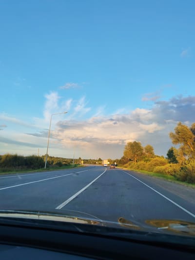 This is a photograph taken from the perspective of a vehicle moving on a highway. The road is asphalt with white lane markings. On either side of the road, there is lush green vegetation and trees. The sky is a vibrant blue with scattered white and gray clouds, some of which are illuminated by the setting sun. There are streetlights visible along the side of the road. In the distance, there are a few vehicles on the highway, including a truck and a car. The overall scene suggests a rural or suburban setting during golden hour. The location is the highway leading into the city of Gornyak, Russia.