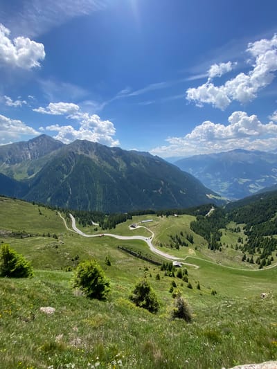 This is a scenic, high-angle, wide shot photograph of a mountainous landscape. The foreground features a grassy slope dotted with small wildflowers and a few scattered evergreen trees. A winding asphalt road with white lines navigates through the mid-ground, with a single white car visible on it. Several small buildings with dark roofs are situated near the road. The mid-ground and background are dominated by rolling green hills and steep, tree-covered mountains, receding into a hazy blue distance. The sky is a vibrant blue, with scattered white, puffy cumulus clouds and thinner, wispy cirrus clouds. The sun is high and bright, creating lens flare in the upper right quadrant of the image. The overall scene depicts a tranquil, natural environment with signs of human infrastructure, likely in a rural alpine setting. The location is identified as St. Leonhard in Passeier - San Leonardo in Passiria, Italy.
