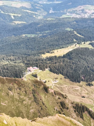 This image provides a high-angle view of a mountainous landscape near Alpnach, Switzerland, focusing on a cable car station and surrounding terrain. In the foreground, a steep, grassy slope descends into a valley. Further down the slope, a cable car station is visible, with a prominent tower supporting cables and several red cable car cabins. The station building itself is a large, red-roofed structure with some adjacent smaller buildings.
Beyond the cable car station, the landscape transitions into a dense forest of evergreen trees that covers the hillsides. Open, grassy clearings with scattered trees and a few buildings are interspersed within the forest. A winding road or path is visible snaking through the landscape. In the far distance, a more spread-out settlement with buildings and infrastructure can be seen on a gentler slope, suggesting a town or village. The sky is clear, indicating a sunny day. The overall scene depicts a popular tourist or recreational area in the Swiss Alps, likely accessible by cable car.