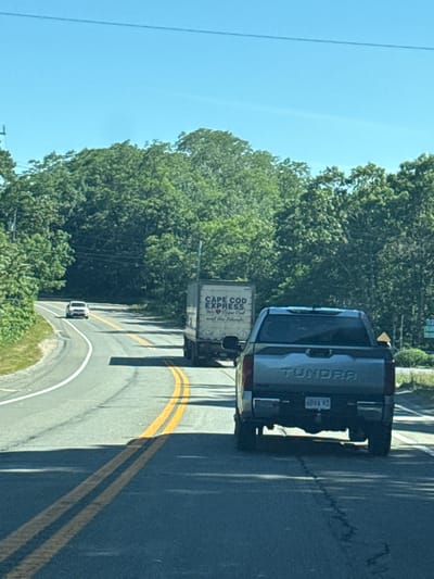 The image shows a two-lane road in Wellfleet, Massachusetts, on a clear day. The road curves to the left, with dense green trees lining both sides. In the foreground, a silver Toyota Tundra pickup truck is traveling in the right lane. The truck has a license plate that reads "68VA 93". Ahead of the Tundra, in the left lane, is a white box truck with "CAPE COD EXPRESS" and a heart logo printed on its side. Further ahead in the left lane, a white sedan is visible. The road surface is asphalt with yellow double lines indicating no passing zones. The sky is bright blue with a thin wire running horizontally across the top of the frame.