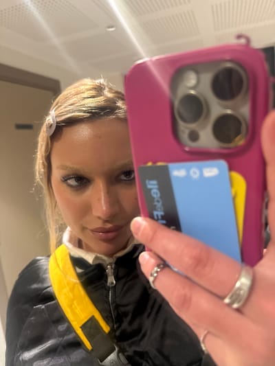 A selfie shows a person with blonde hair, light skin, and multiple rings on their fingers. They are holding a magenta phone case, with a bus pass and a blue card tucked behind. The phone has a triple-lens camera. The person is wearing a black jacket with a yellow accent. The background is a blurred, neutral tone, possibly a wall.