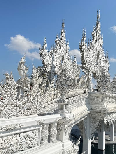 A detailed, ornate white structure featuring elaborate dragon-like figures with flowing, flame-like designs against a bright blue sky with fluffy clouds. The architectural details are intricate and elaborate. The structure appears to be a temple or religious building. The decorations extend upward, creating a complex, ethereal impression. The white color is stark, contrasting with the sky.