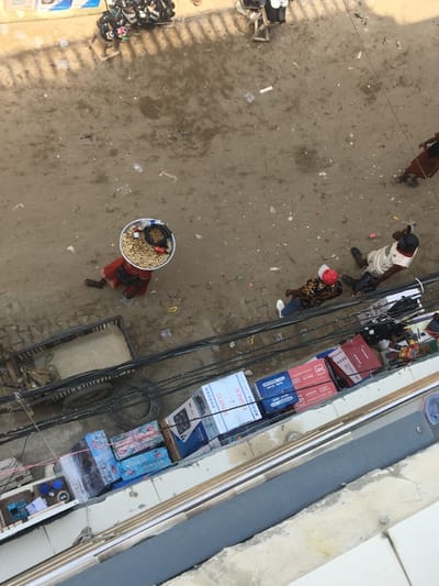 This high-angle image depicts an active street scene in Ojo, Nigeria. The ground is predominantly unpaved dirt, characterized by uneven patches of damp earth and scattered litter, including plastic bottles and wrappers.
In the center-left, an individual in a red garment walks rightward, balancing a large, circular metal tray on their head. The tray contains a substantial quantity of small, light-colored granular items, a black object, and two red cans. To their right, an individual is positioned low on the ground, possibly sitting or resting, wearing a light-colored shirt and a dark baseball cap. Below this person, a third individual stands, clad in a patterned top, dark trousers, and a white baseball cap. Further in the upper-right, the lower torso and legs of a person in a brown garment are partially visible, moving away from the scene's center. In the upper-left, several motorcycles are parked near a small wooden stool.
Multiple black electrical cables span horizontally across the mid-ground. Beneath these cables, along a concrete ledge, numerous cardboard boxes are stacked, some marked with "DJACK" and "M" branding, indicating commercial goods. Adjacent to these boxes on the left are two discarded tires and a dark, rectangular utility box or container. The immediate foreground consists of a concrete building edge or balcony, suggesting the elevated perspective of the photograph.