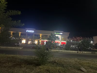 The image depicts a nighttime scene in the city of Kyzgaldak, Uzbekistan, showing a commercial building with illuminated signage. The building features a light-colored facade with several large windows and entrances. Prominent signs include "KARTAL BİLMƏKƏRLƏRİ" with a blue arrow logo, and "NASRİDDİNSAVDO" with a green circular logo. The storefronts display various products, some of which appear to be groceries. Several vehicles are parked in front of the building and on the adjacent road. A few individuals are visible, some standing near the entrance and others near parked cars. The surrounding environment includes trees, grass, and a road. The sky is dark, indicating nighttime.