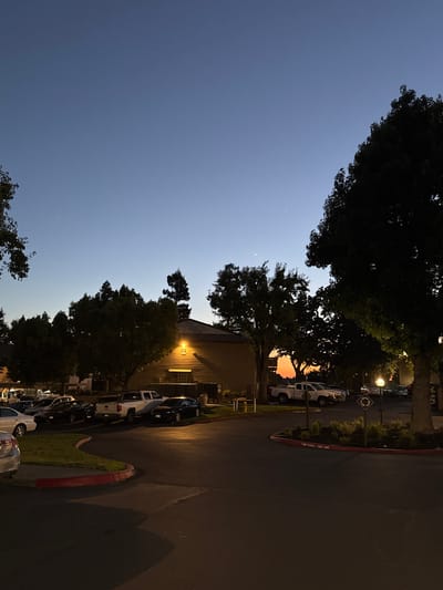 A twilight scene captures a parking lot adjacent to a building with an illuminated exterior light. The sky exhibits a gradient of blues, transitioning to a faint orange glow near the horizon, suggesting a sunset. Several vehicles, including sedans and pickup trucks, are parked in designated spots. Lush green trees, some silhouetted against the sky, frame the scene, with one large tree dominating the right side of the frame. A streetlamp is visible, casting a warm light on the asphalt. A red-curbed island with grass and small plants is present in the foreground. There are no people visible in the image. The overall impression is one of a suburban or commercial area settling into evening.