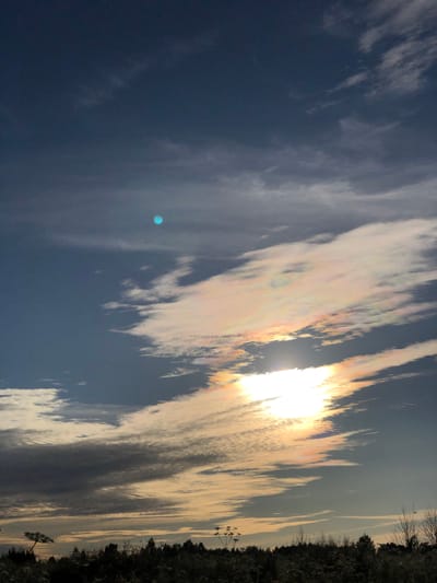 The image shows a sky with scattered clouds at sunset. The sun is partially obscured by clouds, creating a bright, shimmering effect with iridescent colors. A small, distinct blue circle is visible in the upper left portion of the sky. The lower part of the image depicts a silhouetted landscape of trees and vegetation against the twilight sky. There are no people or animals visible in the scene. The location is identified as Staroe Istomino, Russia.