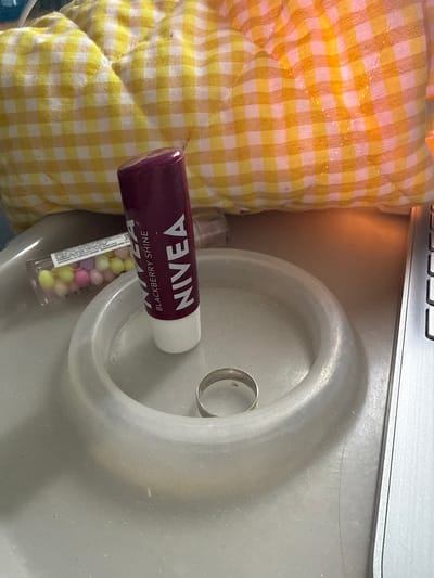 A close-up shot displays a Nivea lip balm tube and a clear plastic container of small, pastel-colored candies on a light gray surface. A silver ring rests inside a shallow, circular dish. In the background, a yellow and white checkered fabric is visible. The scene is illuminated by warm, artificial light, casting orange hues on the background fabric and creating a soft glow on the surface. The image does not contain information about the city of Nerudstal, Ukraine.