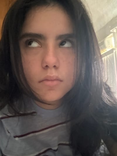 The image displays a close-up portrait of a single individual, centered in the frame. The subject has long, dark, wavy hair and a light to medium skin tone with visible freckles on the nose and cheeks. They are wearing a light grey or blue short-sleeved shirt with alternating thin white/light grey and dark red/maroon horizontal stripes. The individual's eyes are directed upwards and slightly inward, while the mouth maintains a neutral, closed expression. The background is out of focus, revealing an indoor setting. A bright, rectangular light source is present in the upper right background, illuminating a portion of a light-colored, possibly textured wall. There are no visual cues within the image to identify the specific location in the city of Santiago, Mexico.