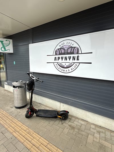 An electric scooter is parked on a sidewalk in front of a building with a large sign. The scooter is black with red accents and has a black bag attached to the handlebars. To the left of the scooter is a silver trash can. The building has dark gray siding and a white sign with black lettering and graphics. The sign appears to be an advertisement. The sidewalk is paved with gray and yellow rectangular tiles. A portion of a green sign with a white symbol is visible on the left side of the building. The weather appears to be overcast.