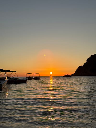 The image captures a sunset over the Ionian Sea in Palaiokastritsa, Greece. The sun, a brilliant orange orb, is positioned centrally on the horizon, casting a distinct, shimmering vertical reflection across the water surface towards the foreground. The sky transitions from a light blue-gray at the top to vibrant orange and yellow hues near the horizon. Two circular lens flares, one prominent, are visible in the upper-left quadrant.

In the middle-ground, four small motorboats are anchored in the calm, rippled water. The boats are rendered in silhouette, each featuring a dark, open canopy structure. The leftmost boat is partially visible. To the far right, a large, dark, rocky landmass or cliff rises steeply from the water, silhouetted against the bright sky. Smaller dark rock formations are also visible in the water near the base of this main landmass and further to its left. No individuals or specific actions are discernible within the frame.