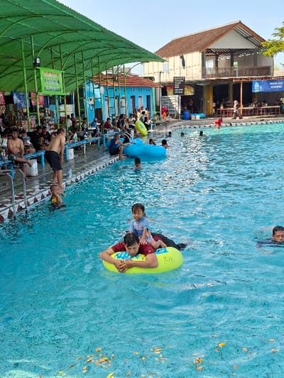 The image shows a swimming pool located in the city of Kecamatan Tulung, Indonesia, at coordinates -7.592 latitude and 110.649 longitude. The pool is surrounded by a deck with several people sitting and standing around, including a group of people in the background sitting at a table. In the pool, there are several people swimming and playing. Two people, a man and a young child, are riding a yellow inflatable ring. There is a blue inflatable ring in the background, not being used. The water in the pool is blue and rippled. There are leaves on the surface of the water and the water appears to be shallow, revealing the pool bottom. The scene is set against a backdrop of a building with a green roof, palm trees, and blue sky. The building appears to be part of the swimming pool complex.