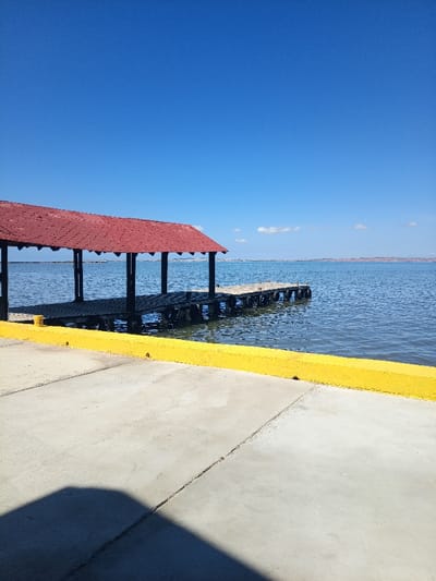A pier with a red roof extends over a body of water under a clear blue sky. Black support posts and tires are visible beneath the pier. A yellow curb borders a gray concrete surface in the foreground. The water appears calm, and land is visible on the distant horizon. A dark shadow is cast on the concrete.