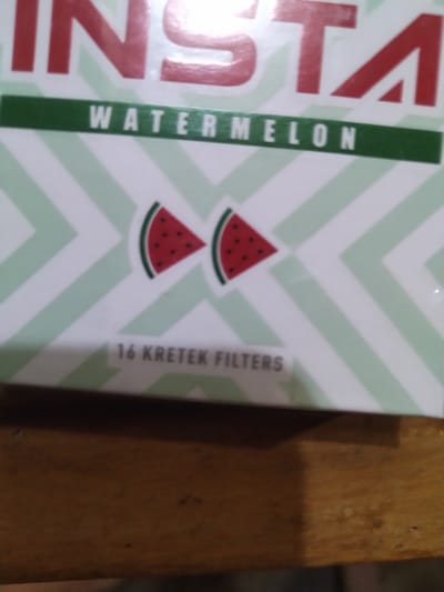 A close-up shot shows a retail product, likely a pack of cigarette filters, resting on a wooden surface. The product packaging is primarily white with light green diagonal stripes. Prominently displayed on the packaging is the red text "INSTA" at the top. Below this, a green band contains the white text "WATERMELON". Two stylized watermelon slices are depicted in the center of the packaging. Near the bottom of the packaging, white text reads "16 KRETEK FILTERS". The wooden surface appears to be a table or shelf, showing grain and some minor imperfections. The image is taken from a slightly elevated angle, focusing on the product. The location in Gampongbaro, Indonesia is not discernible from the provided image.