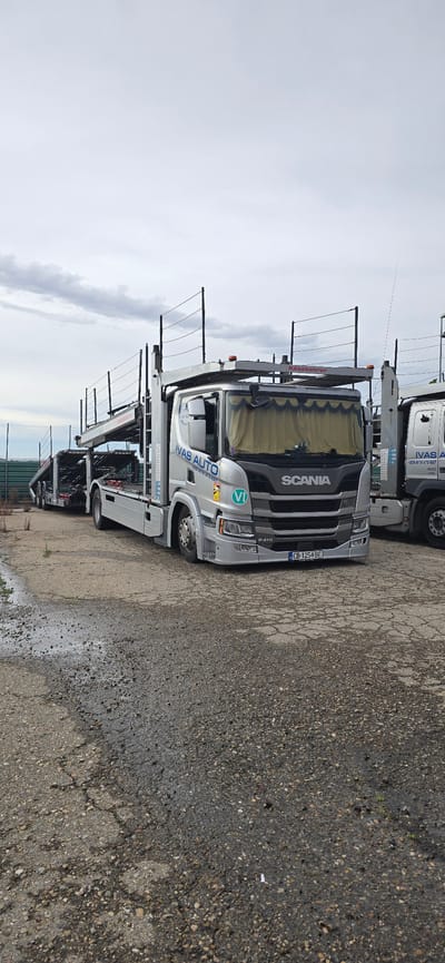 This is a vertically oriented, eye-level, outdoor photograph of a Scania car carrier truck parked on a wet, cracked asphalt lot. The truck is silver and grey, with "SCANIA" prominently displayed on the front grille. The license plate reads "CB-1254-BE". The truck has a car-carrying apparatus attached, with several rows of metal supports visible. The cab of the truck features the text "VAS AUTO" and a circular green and white logo with the letter "VI" inside. Another truck, also a car carrier, is partially visible behind the main truck, with "IVAS A" and a phone number "+359 878 879 637" visible on its side. The sky is overcast and appears grey, suggesting a cloudy or potentially rainy day. The ground is wet, with puddles and reflections visible, indicating recent precipitation. The overall scene appears to be a commercial or industrial setting, possibly a parking lot for a transport company, given the nature of the vehicles. There are no people visible in the frame. The lighting is dim, consistent with an overcast sky.