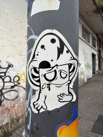 A sticker, primarily white with black cartoon elements, is affixed to a gray metal pole. The design features a whimsical, perhaps skeletal, figure with outstretched hands. One half of the head has a lightning bolt design, while the other features circular spots. A yellow smiley face sticker is partially visible adjacent to the main design. The pole shows signs of wear and tear. Graffiti is visible on a wall or surface behind and below the pole.