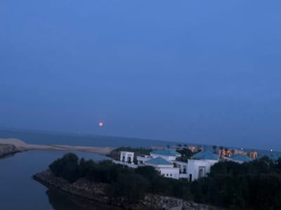 The image depicts a coastal scene at dusk or early evening in Fnideq, Morocco. A body of water, possibly a lagoon or river estuary, is visible in the foreground, with dark water reflecting the dim light. A sandy beach or dune separates the water from the ocean.
In the middle ground, a complex of white buildings with distinctive turquoise-colored roofs sits atop a rise. These structures appear to be part of a resort or residential development. Trees and shrubbery obscure the base of the buildings.
In the background, the ocean stretches to the horizon, where a faint, reddish orb (likely the moon) is visible. The sky is a deep blue, suggesting the waning daylight. No people are visible in the image. The scene is still and serene.
