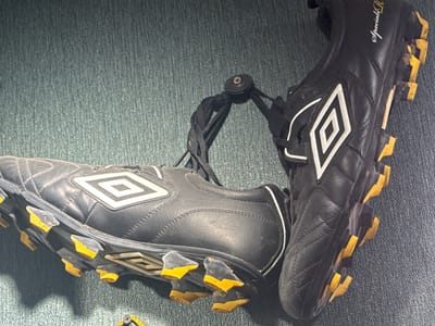 A pair of black Umbro soccer cleats with yellow accents is positioned on a textured, dark green surface. The cleats are connected by their laces, which are tied together in the center with a small, black circular clasp. The left cleat is lying on its side, revealing the sole with its distinctive yellow studs. The right cleat is upright, showing the side and laces. The surface appears to be a rug or carpet. The scene is illuminated from above, casting shadows to the left of the cleats. The cleats appear to be used, with some dirt visible on the soles and studs. The background is out of focus, but it appears to be a continuation of the same textured surface. The context of Protasovo, Russia, does not appear to be directly represented in the visual elements of the image itself.