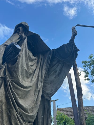 The image captures a low-angle view of a statue, possibly depicting a cloaked figure holding a tool. The statue appears to be made of a weathered, gray material and is draped with fabric that creates deep folds. In its left hand, the figure holds a cylindrical object with a textured surface. The right arm is raised, and the hand grasps the top of a long-handled tool, possibly a scythe or hoe, with a curved blade. The statue is supported by wooden poles.
In the background, a bright blue sky with scattered white clouds dominates the upper portion of the image. Below the sky, partially visible are residential buildings with tiled roofs and green foliage. The context of Prokhladny, Russia is provided as the location. The overall impression is of an outdoor setting, potentially a garden or a public space, where the statue is either being displayed or is under construction/restoration.