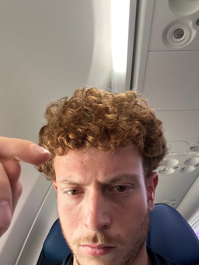 A man with light skin and curly brown hair is in an airplane. He is giving a thumbs up. Overhead compartment features are visible. The location is unknown and there is no indication the photo was taken in Cleveland, United States of America.