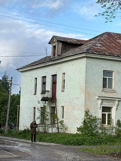 A medium outdoor shot captures a three-story building in Ulyanovka, Russia, with a dilapidated exterior. The building, with a rusted brown roof, has light-blue peeling paint. Various-sized windows are apparent. A metal balcony protrudes from the second floor, accessible via a red door and a metal ladder. Overgrown vegetation partially obscures the lower right of the facade. A person is walking in front of the building. A utility pole is visible on the left side and trees are visible along the bottom of the picture. The sky is overcast with visible clouds.