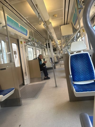The image depicts the interior of a brightly lit İZBAN commuter train car, likely operating within Gaziemir, İzmir, Turkey. The car features rows of white-backed seats with blue patterned fabric, many of which are vacant. Silver-colored vertical support poles and horizontal handrails with hanging loops are visible throughout the cabin. Beige and white panels line the walls and ceiling, which incorporates overhead lighting. An overhead display on the left side shows a route map, characteristic of the İZBAN system.

One adult male is seated on the left side of the train, approximately midway down the car. He has dark hair and is wearing a dark jacket, dark trousers, and dark shoes. He is holding a white mobile phone with both hands, his gaze directed downwards towards the device. The floor is light-colored, with grey textured matting present near the doorways. Windows along the side of the train show a blurred, indistinct exterior environment.