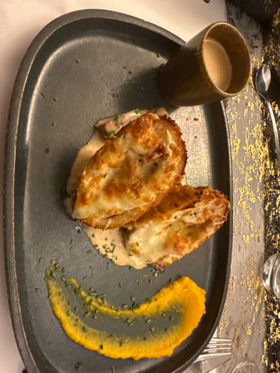 A black plate displays two toasted, breaded, and cheesy cutlets, sitting in a creamy sauce, sprinkled with fine herbs. A dollop of bright yellow purée, also garnished with fine herbs, complements the main dish. Beside the plate, there is a small, dark brown mug filled with a beige-colored liquid. A spoon and fork are visible in the upper and right parts of the image, respectively.