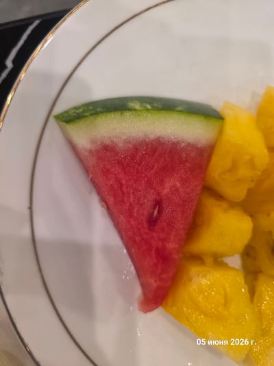 The image displays a close-up, top-down view of a white ceramic plate containing cut fruit, located in the city of La Louvière, Belgium. On the left, a triangular slice of watermelon presents a gradient from dark green rind to a narrow band of white pith, transitioning into vibrant red flesh. A single, small, elongated white seed is visible within the red flesh. To the right, several irregularly cut, bright yellow fruit chunks, consistent with either pineapple or mango, are arranged. The white plate features two concentric gold-colored decorative rims. A dark, striped surface is partially visible in the upper left background. The date "05 июня 2026 г." (05 June 2026) is overlaid in white text at the bottom right of the image. No people are visible.