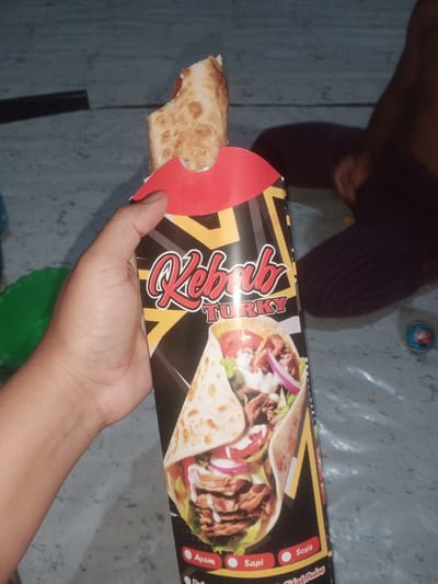 A person's hand holds a kebab in a red and black cardboard wrapper, featuring an image of a kebab on the front. The wrapper has "Kebab Turky" written in large red lettering with a yellow outline. The kebab itself appears to be a wrap containing meat, vegetables, and sauce. The background is a gray, textured surface, possibly a floor or ground, with a blurred dark purple shape in the upper right corner, which appears to be a person sitting. A green circular object is visible on the left. The image is taken from a slightly elevated, close-up perspective. The location is specified as Cotseutui, Indonesia, although this context is not visually evident in the image itself.