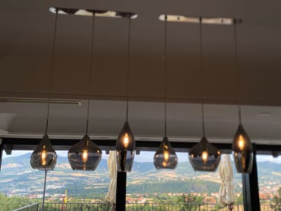 The image shows a row of six pendant lights with smoky glass teardrop shades, each containing a lit Edison-style bulb. The lights are suspended from a rectangular ceiling fixture. The background is a panoramic view of a valley with rolling hills, vineyards, and scattered buildings, indicating a location outside of a dense urban area. The lighting suggests it is daytime, possibly late afternoon given the warm glow. The scene appears to be taken from an indoor vantage point looking out through a large window or opening. The architecture of the room is modern and understated, with a neutral-colored ceiling and walls, which allows the lights and the exterior view to be the main focus. The specific location is identified as a restaurant or residential property in the Arandjelovac Municipality, Serbia, which is known for its natural beauty and wine-producing regions.