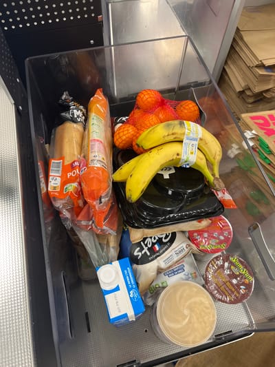 A plastic bin filled with groceries sits atop a stainless steel surface in a grocery store. The bin is filled with a variety of items, including a bag of oranges, two baguettes, three bananas, two containers of yogurt, a container of hummus, and a container of cereal. The items are arranged in a haphazard manner, with some spilling out of their containers. The bin is sitting on a stainless steel surface, and the store is sparsely populated. The location is based on coordinates (latitude: 49.283, longitude: 3.999) and is the city of Saint-Brice-Courcelles, France. The store is likely a local supermarket.