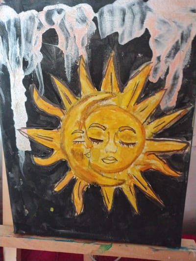 This is a close-up, eye-level shot of a painting on canvas. The painting depicts a stylized, bright yellow sun with a distressed, childlike face. The sun's rays are sharp and pointed, painted in shades of yellow and orange with black outlines. Its face has two large, crescent-shaped eyes, a small nose, and a wide, smiling mouth with two visible teeth. The sun is set against a black background, which is partially obscured by abstract, cloudy shapes painted in white and peachy-pink hues on the left side of the canvas. These clouds appear to be swirling and possibly represent smoke or celestial phenomena. In the lower part of the painting, there are hints of white and grey textures that might suggest a landscape or foreground elements. A small, bright yellow dot is visible in the lower right portion of the black background. The painting is displayed on an easel, with the wooden supports of the easel visible on the right side of the frame. The lighting in the scene is bright, highlighting the colors and textures of the painting. The overall mood of the artwork is energetic and possibly a bit melancholic due to the sun's expression, but the vibrant colors suggest a sense of resilience or hope. There is no visible text in the image.