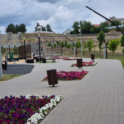 The image depicts a public park in the city of Liski, Russia, on a cloudy day. The foreground features a paved walkway lined with flower beds containing a variety of colorful petunias in shades of pink, purple, and white. Two dark brown, slatted trash receptacles are visible along the walkway.

In the midground, more flower beds are present, along with wooden park benches and trash receptacles. A children's playground area with a green toy truck and black rubber matting is partially visible on the left. Further back, a set of concrete stairs ascends a gentle slope.

The background shows a white gazebo on top of the slope, surrounded by trees. A residential building and more trees are visible on the horizon. Several people are scattered throughout the scene, walking on the stairs and in the park area. The overall impression is that of a well-maintained urban green space.