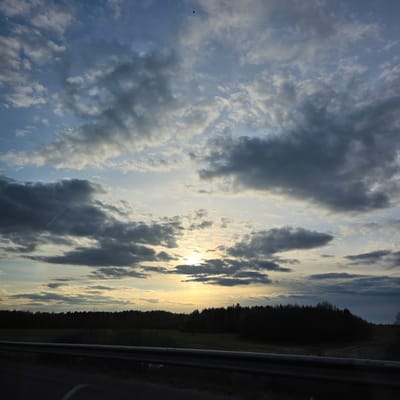 The image displays a serene landscape during either sunrise or sunset, characterized by a dynamic sky and a dark forest silhouette. The upper portion of the frame is filled with a vast sky, showcasing a mix of bright blue, soft white, and darker grey clouds. The sun, partially hidden by clouds, emits a warm, golden glow from the right side, casting visible rays of light through the cloud formations.
Along the horizon, a dense line of dark trees forms a stark silhouette against the bright sky, suggesting a thick forest or woodland. Below the tree line, a strip of lighter-toned field or grass is discernible. In the foreground on the right, a metallic guardrail runs horizontally, indicating the presence of a road or highway adjacent to the natural environment. A small, dark, bird-like speck is visible high in the left part of the sky. The scene evokes a tranquil atmosphere in the rural setting of Талачынскі сельскі Савет, Belarus. There is no visible text in the image.