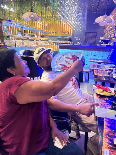In a dimly lit restaurant with a lively ambiance, a woman and a man are seated at a table. The woman, with dark hair and wearing a maroon top, is looking to her left, her mouth open as if speaking. The man, wearing a white Vans baseball cap and a white t-shirt, is leaning forward, holding a fan adorned with a red and white design. He appears to be looking at the fan, his eyes closed. The restaurant's interior features a buffet-style display with covered food containers, wooden accent walls with vertical illumination, and a ceiling adorned with hanging plants and decorative lights. A bar area with a blue, patterned backdrop and overhead shelves with glassware is also visible. On their table, there are plates of watermelon and a glass with a Pepsi logo. The overall scene captures a moment of interaction between the two individuals in a vibrant dining setting.