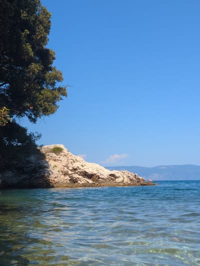 The image depicts a serene coastal scene under a clear blue sky. In the foreground, shallow, transparent water with ripples reveals the seabed below. To the left, the edge of a large, dark green tree overhangs the water. In the mid-ground, a rocky island with a light-colored, textured surface juts out of the sea. A small patch of green vegetation is visible on the island. In the background, a calm, deep blue sea stretches to the horizon, where hazy, blue mountains form a backdrop under the expansive sky. A small orange kayak with a person in it is visible on the water near the rocky island. The overall impression is one of tranquility and natural beauty, likely a coastal area within or near the city of Labin, Croatia, given the characteristic Adriatic landscape.