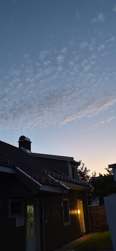 The image depicts the twilight sky over a residential building. The sky is a gradient of blue and pale orange, with scattered cirrus clouds. The building in the foreground is a two-story house with a dark tiled roof and green siding. A chimney with a silver cap is visible on the roof. A light fixture above a doorway illuminates the entrance and a small patch of lawn. The scene appears to be in a suburban area. The image does not provide specific clues to pinpoint the location within Lillehammer, Norway. There are no people or animals visible in the image.