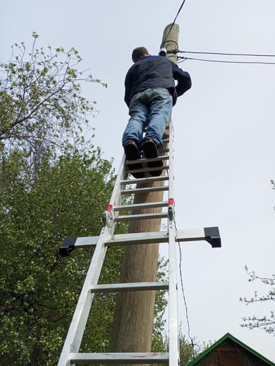 A man is climbing an aluminum ladder leaning against a wooden utility pole. The ladder has a support bracket on one side with a black rubber foot. The man wears a dark blue jacket, blue jeans, and brown shoes. Electrical wires are connected to the top of the pole. Trees with budding leaves are visible on the left side of the frame. A glimpse of a green-roofed structure appears at the bottom right. The sky is overcast. The location is Монастырка, Russia.