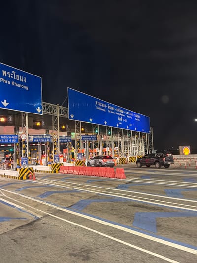 The image shows a nighttime view of a toll plaza in Bang Kaeo Subdistrict, Thailand. The toll plaza has multiple lanes, each marked with signs indicating directions and lane numbers. Several cars are visible, including a silver sedan and a dark-colored pickup truck, some of which are passing through the toll booths. The toll booths themselves are brightly lit with yellow and red lights, and there are barriers and cones to guide traffic. The background shows a dark sky with some faint clouds. Signs above the lanes provide information in both Thai and English, indicating destinations such as "Phra Khanong" and "Dao Khanong." The ground is asphalt, with painted lane markings and some blue-colored areas.