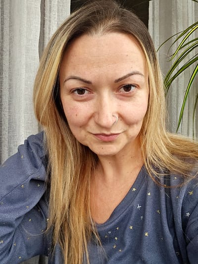 A head-and-shoulders portrait of an adult female positioned centrally. She has long, light blonde hair with visible darker roots, brown eyes, and fair skin, with a small, studded, circular nose ring visible in her right nostril. She wears a ribbed, V-neck, long-sleeved top in a slate blue or grey hue, embellished with small, scattered metallic gold and silver star-shaped patterns. The individual looks directly forward with a neutral to slight smiling expression. The background consists of light-colored, vertically pleated fabric, consistent with curtains or drapes, visible on both sides of the subject. A portion of a green and yellow striped houseplant leaf is visible behind her right shoulder. The image provides no discernible information to suggest a specific location within the city of Leicester, United Kingdom, or any other geographical area.