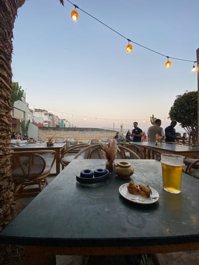 This image depicts an outdoor seating area of a restaurant or cafe, likely located in Taghazout, Morocco, given the coastal architecture and vegetation visible in the background. The time of day appears to be dusk or early evening, with string lights illuminating the scene.
In the foreground, a dark-colored table is set with two blue items, a small golden pot with dried flowers, and a plate with two small food items. To the right of these objects is a tall glass filled with a golden-amber colored beverage, likely beer. The table has a textured, possibly stone or metal, surface. Two chairs with woven backs and seats are visible to the left and right of the table. A portion of a palm tree trunk is on the far left of the frame.
In the middle ground, other tables and chairs are arranged. Some patrons are seated on sofas and chairs in the background, appearing to be socializing. Several individuals, presumably staff, are standing and interacting with patrons or each other. The environment suggests a relaxed, social atmosphere.
The background reveals a street or pathway with buildings characteristic of a coastal Moroccan town, including multi-story structures and balconies. The sky is a pale gradient of blue and orange, indicating sunset. Cacti and other arid-climate plants are visible, consistent with the region. The overall scene conveys a pleasant, social dining or drinking experience in an outdoor setting.
