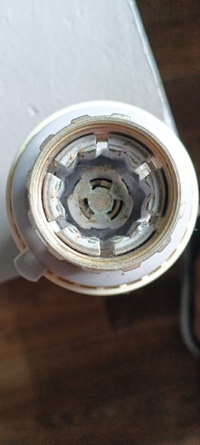 A close-up, overhead view shows the interior of a white and brass colored electrical socket. The socket has a threaded brass fitting with six metal contacts that appear to be slightly corroded and dusty. These contacts are spaced evenly around a central circular opening, revealing a dark interior. The white outer casing has a slightly textured surface. The background is a blurred mix of white and dark brown surfaces, suggesting an indoor environment with a desk or table and possibly a wall. There are no people or actions depicted in the image. The location within the city of Tecnimont camp, Russia, is not visually represented in the image.