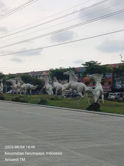 A daylight scene captured on 2025/08/04 at 14:48 depicts an urban environment in Pusakarakyat, Kecamatan Tarumajaya, Indonesia. The foreground features a wide, light-grey paved road surface, appearing dry and clear. Beyond the road, a raised, manicured grassy median displays a prominent art installation consisting of approximately eight large, white, stylized horse statues. Each statue is depicted in various dynamic running or galloping poses, with detailed manes and tails suggesting motion. Irregular dark rocks or boulders are placed near the base of some statues on the green grass. In the midground and background, a line of dense green trees partially obscures multi-story buildings with varied facades, including red, orange, and blue sections, suggesting commercial or mixed-use structures. Several light-colored vehicles, including white vans and dark cars, are visible on a road or parking area behind the statues. The sky overhead is overcast with visible grey and white clouds. Multiple black utility lines stretch horizontally across the upper portion of the frame. No individuals are visibly present in the scene.
