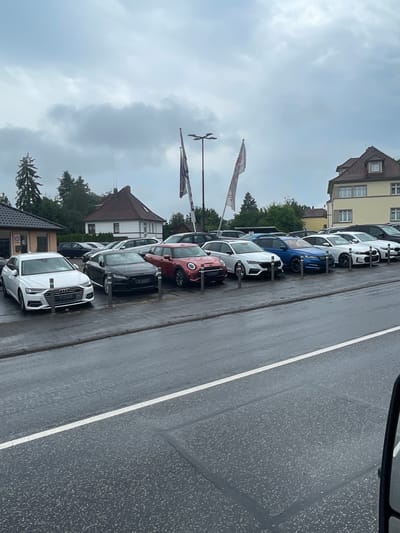 The image depicts a car dealership lot in Lauterbach, Germany, under a cloudy sky. A variety of new cars are parked in neat rows on a wet asphalt surface. In the foreground, a white Audi A6 Avant is prominently displayed, followed by a black Audi TT. Further back, a red Mini Cooper, a white Skoda Octavia Combi, and a blue Skoda Superb are visible. Several other vehicles, including white BMWs and a dark SUV, are parked in the background. Buildings, including a dealership office and residential houses, are situated behind the car lot. Flags are displayed on poles, and trees and streetlights are also present. The overall scene suggests a typical day at a car dealership, possibly experiencing light rain given the wet road surface.