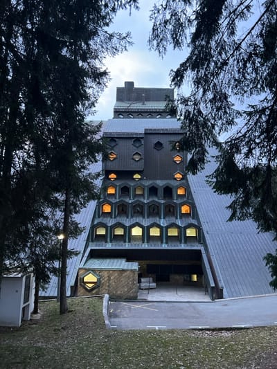 The image displays a large, geometrically complex building situated in Kolašin, Montenegro, framed by dense evergreen trees under an overcast sky. The multi-tiered structure features steep, dark metal-paneled roofs and facades, creating a stepped, almost pyramid-like silhouette. The building's exterior incorporates distinct window patterns: hexagonal windows are visible on the upper levels, while multiple lower tiers display rows of uniformly shaped, hooded rectangular windows. Many interior lights are active, casting warm yellow and bright white illumination from these windows. An asphalt driveway, marked with white lines including an 'X' symbol, leads to a recessed entrance area at the base of the building. A single streetlamp illuminates the left side of the driveway, adjacent to a grassy embankment where a white utility box is also present. No persons or vehicles are visible in the scene.