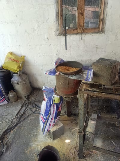 The image depicts a cluttered storage area or workshop, possibly in the city of Auly, Ukraine. The scene is indoors, with a rough, whitewashed brick wall forming the background. A wooden-framed window with a dirty, multi-paned glass is visible in the upper right portion of the image. The floor is concrete and littered with scattered grain, dust, and what appears to be animal feed.
Several objects are present, suggesting agricultural or feed processing activities. To the left, there are two large, dark cylindrical containers and a silver metal milk can. A yellow plastic bag with Cyrillic lettering, possibly "АВРОРА МУЛЬТИМАРКЕТ," is leaning against the milk can. A small, light brown ceramic jug sits on the floor near the base of the milk can.
In the center of the image, a large, rusty red metal drum is positioned under a wide, dark metal bowl filled with corn kernels. The drum appears to be part of some machinery. To the right of the drum, a rough, weathered grey stone block rests on a rusty metal structure that resembles a workbench or processing platform. Bags of what appears to be animal feed are also visible, some with Cyrillic lettering. One large white and blue bag is prominent in the foreground, with text that includes "РОЙЛЕРІВ" and "BEST MIX."
Electrical cords and a rope are strewn across the floor. In the lower right foreground, part of a dark bucket or container is visible. The lighting suggests an indoor environment with some ambient light entering from the window or an unseen source. There are no people visible in the image. The overall impression is one of a working space that is utilitarian and somewhat disorganized.