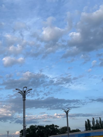 The media file shows a cloudy sky with a light blue hue. Dark gray and white clouds are scattered across the sky, with some areas appearing more illuminated than others, suggesting the presence of sunlight breaking through. Two modern-style streetlights with distinctive, curved arms are visible, one closer to the foreground and another further in the distance. The streetlights are silver and appear to be made of metal. To the right of the frame, a row of dark green trees and taller, thin trees, possibly poplars, are silhouetted against the sky. The background also shows hints of a blue and yellow structure, possibly a building or fence, on the far right. The overall scene suggests an outdoor setting, potentially a street or park, during the daytime, possibly late afternoon or early evening due to the cloud cover and soft lighting. There is no visible text.