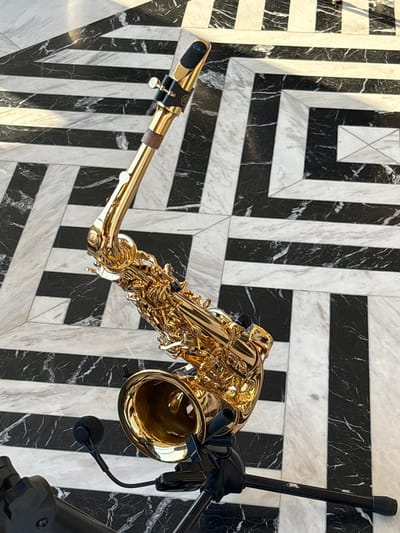 A golden saxophone is placed on a black and white striped marble floor. The saxophone is angled, with its bell facing downwards and towards the left. A small black microphone is attached to the bell of the saxophone with a flexible arm. The saxophone rests on a black stand. The floor has a geometric pattern of alternating black and white marble stripes, with some sections featuring a marbled texture within the black and white areas. The lighting suggests an indoor setting, possibly a stage or a performance area, given the presence of the microphone and stand. The scene is focused on the saxophone and its immediate surroundings, with no people visible. The location is identified as Kepsha, Russia.