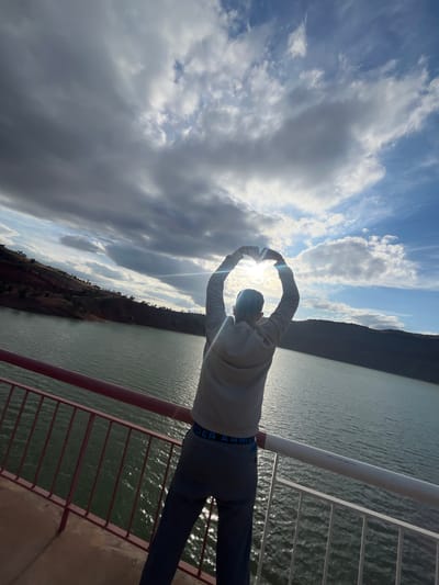 A rotated, medium shot shows a person standing on a structure overlooking a body of water, likely the Barrage Ait Ouarda, near Tabaroucht, Morocco. The person has their arms raised, forming a circle with their hands around the sun, which is partially obscured by clouds. The person is wearing a light-colored long-sleeved shirt, dark pants with a visible "UNDER ARMOUR" waistband, and a hat or hood. The water is reflecting sunlight, creating a shimmering effect. The structure the person is standing on appears to be a bridge or dam, with a metal railing visible in the foreground. In the background, there are hills or mountains. The sky is partly cloudy, with the sun breaking through. The entire image is rotated counterclockwise.