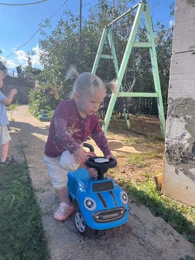 A young blonde girl with two pigtails, wearing a purple shirt with a deer design and white pants, sits in a blue toy car. She is holding the steering wheel and looking to her left. Her feet are in pink sandals. The car has black accents and "HZ SPEEDRUNNER" written on the side. The scene is outdoors, with a path made of concrete slabs and dirt, bordered by green grass and some leafy bushes. In the background, there is a green metal structure, a tree with red berries, and a blue sky with white clouds. Another child, wearing a light blue shirt and striped shorts, is visible in the lower part of the frame, with their back to the camera. The image appears to be taken in daylight, suggesting a sunny day. The setting looks like a backyard or outdoor play area in Rakhinka, Russia.